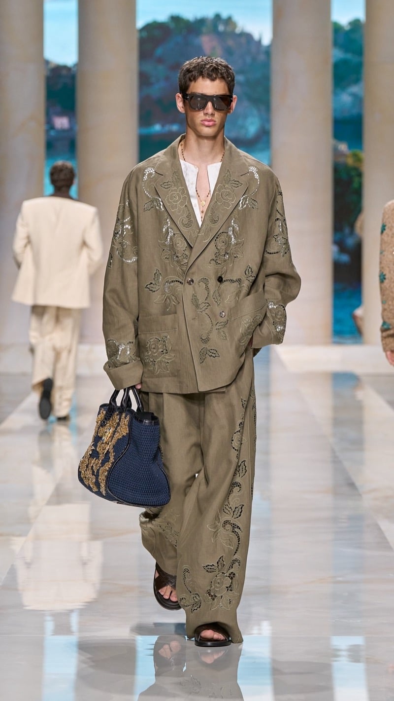 Model in flat-top sunglasses wears an oversized olive linen suit covered in cutwork embroidery and metallic floral applique, layered over a henley, paired with dark sandals and a deep blue and gold beaded tote