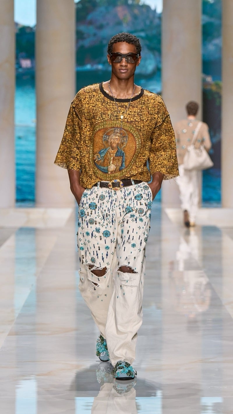 Model in sunglasses wears an oversized golden mosaic-print tee depicting a Byzantine religious icon, paired with light wash distressed jeans densely beaded with turquoise floral motifs and jeweled embroidered shoes