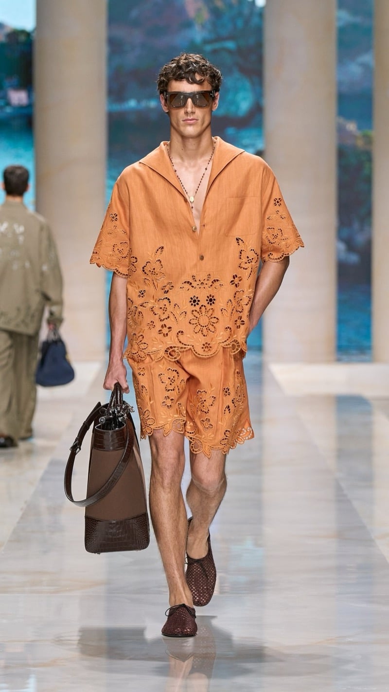 Model in shield sunglasses wears a relaxed orange linen sailor-collar short-sleeve shirt and matching shorts set with scalloped cutwork embroidery, paired with dark crochet derbies and a tan and dark croc-embossed tote