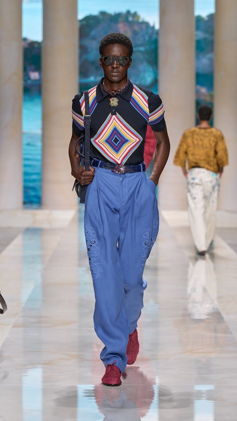 Model in shield sunglasses wears a ribbed short-sleeve knit polo with a multicolored Aztec diamond motif, tucked into wide pleated chambray blue linen trousers with floral cutwork embroidery, paired with red crochet shoes and a deep blue bag