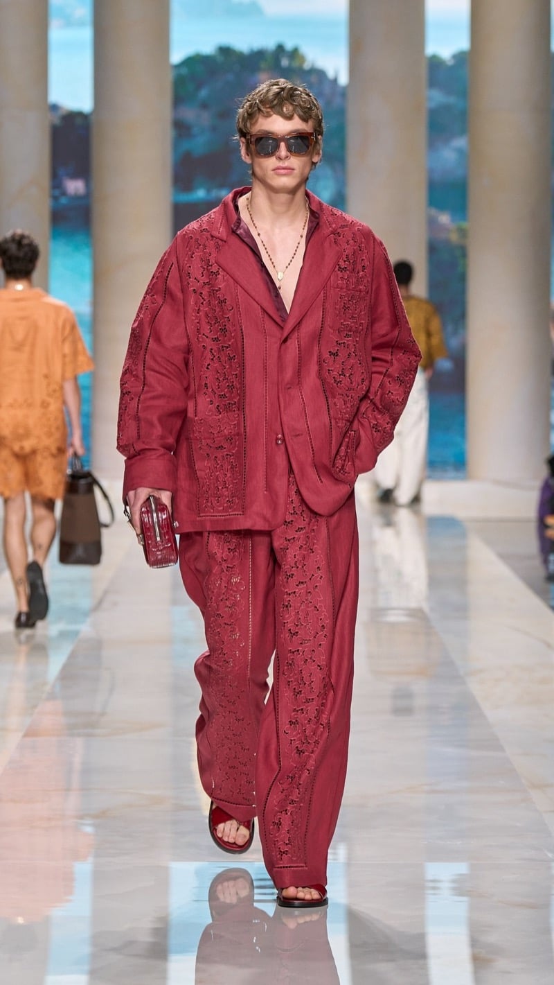 Model in tortoise sunglasses wears an oversized deep red linen set densely covered in tonal beaded floral cutwork embroidery, layered over a layered open shirt, paired with red slide sandals and a small croc-embossed clutch