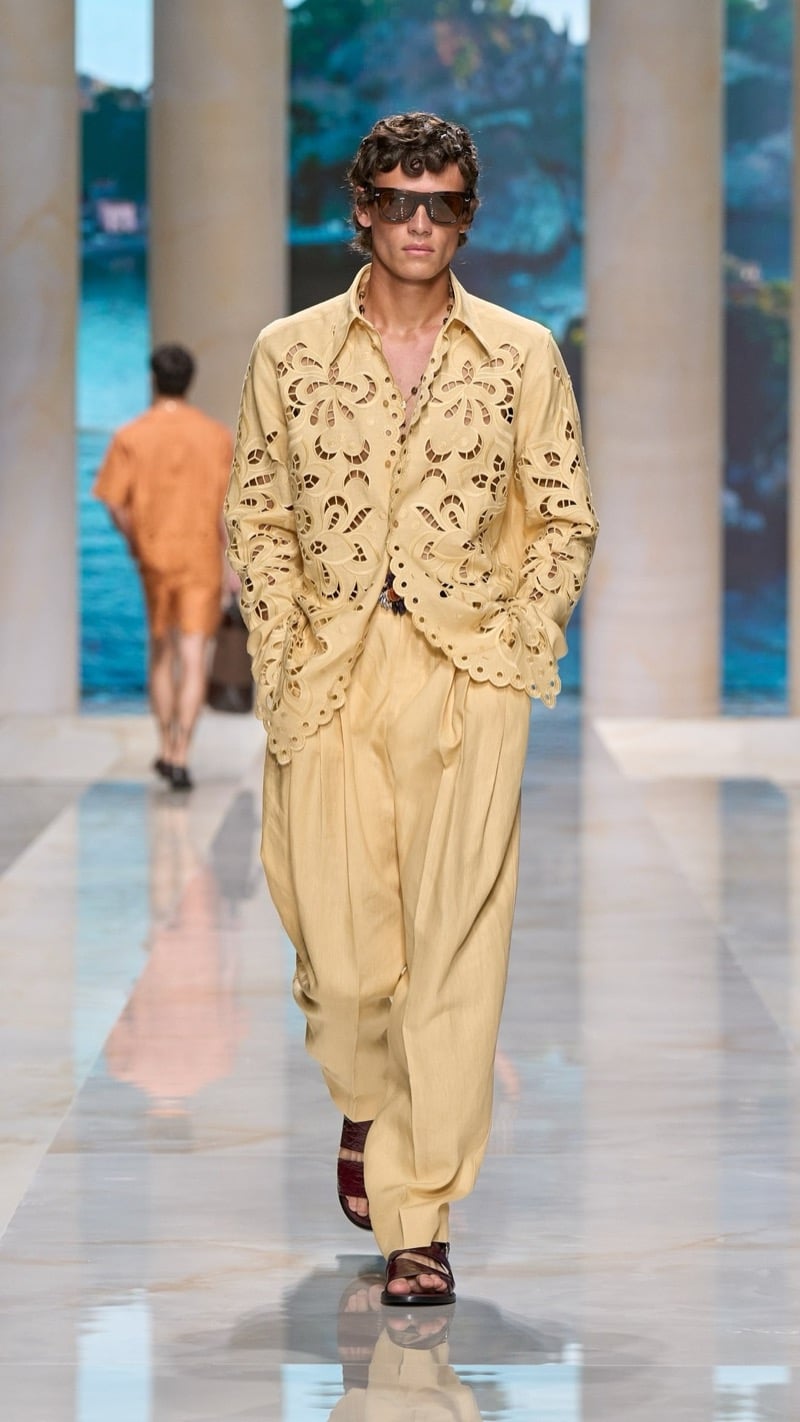 Model in shield sunglasses wears a relaxed pale yellow linen shirt with elaborate floral cutwork embroidery, tucked into wide pleated pale yellow drawstring trousers