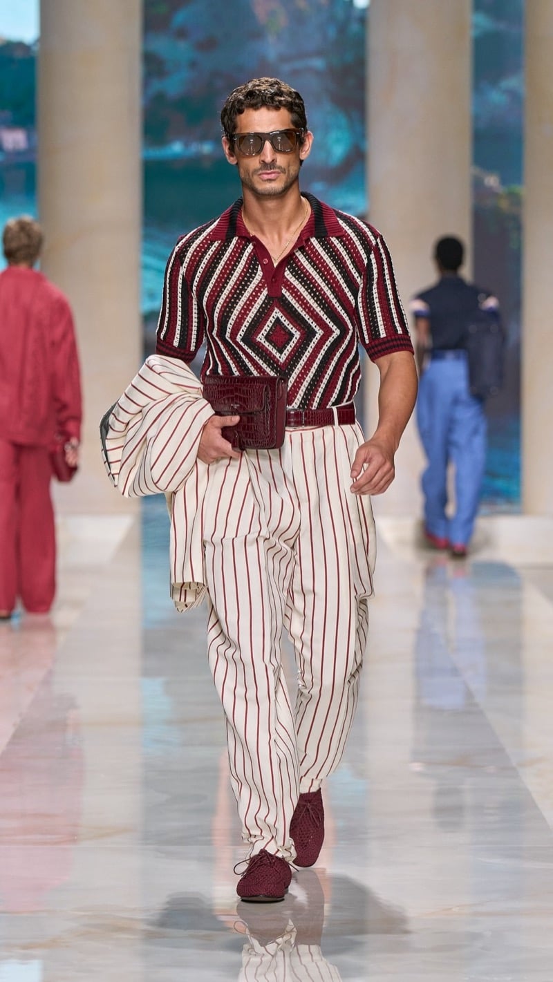 Model in shield sunglasses wears an openwork knit polo with a deep red, ivory, and dark Aztec diamond motif, tucked into wide pleated awning-striped trousers with a croc clutch tucked at the waist, paired with red crochet derbies