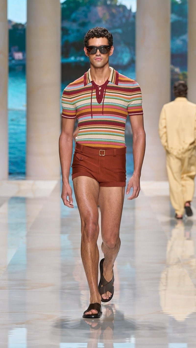 Model in sunglasses wears a multicolored striped short-sleeve knit polo with a laced placket, tucked into deep rust micro shorts with a buckled belt, paired with brown sandals