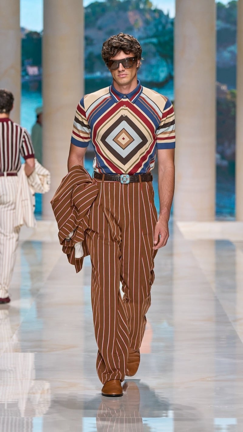 Model in shield sunglasses wears a short-sleeve knit polo with a multicolored Aztec diamond motif in tan, deep red, and blue, paired with wide pleated rust pinstripe trousers, an ornate buckled belt, and tan loafers