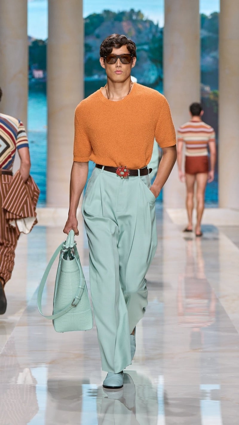 Model in tortoise sunglasses wears a relaxed orange short-sleeve knit crewneck, tucked into wide pleated mint linen trousers with a jeweled buckle belt, paired with mint loafers and a mint croc-embossed top-handle bag