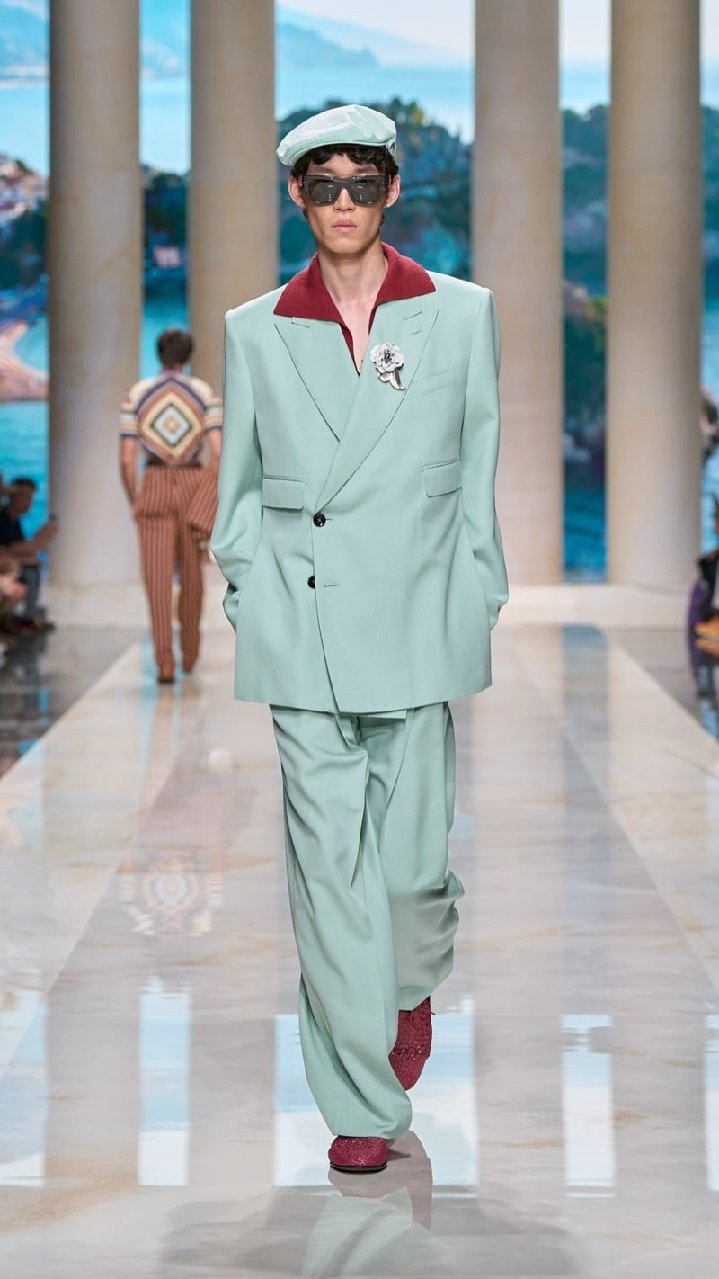 Model in a flat cap and shield sunglasses wears an oversized mint linen suit with peaked lapels and a jeweled brooch, layered over a deep red polo, paired with wide pleated trousers and red crochet shoes