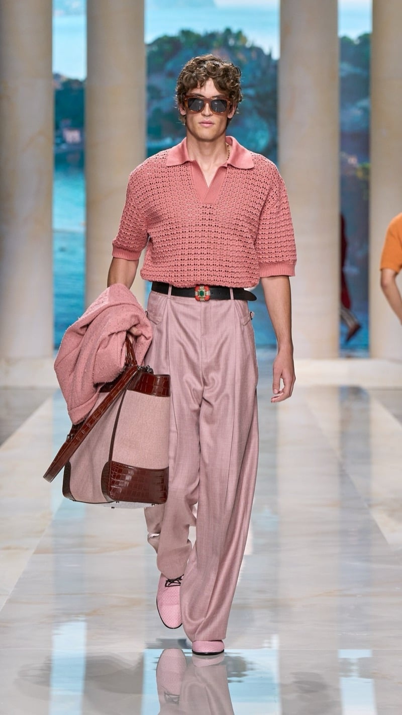Model in tortoise sunglasses wears a short-sleeve pink openwork knit polo, tucked into wide pleated dusty pink trousers with a jeweled buckle belt, paired with pink shoes and a pink suede and brown croc-embossed weekender bag