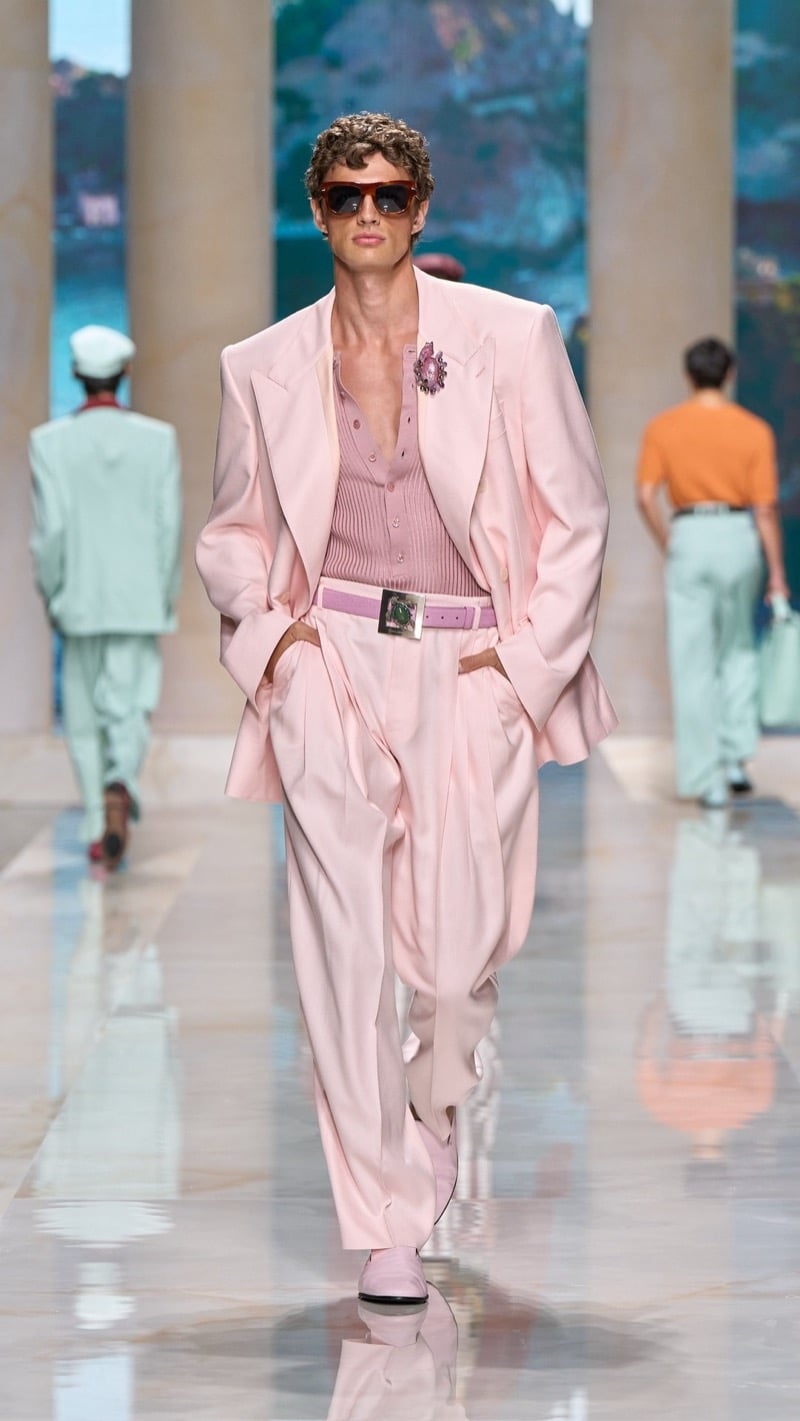 Model in tortoise sunglasses wears an oversized pale pink linen suit with peaked lapels and a jeweled brooch, layered over a deeper pink ribbed knit henley, paired with wide pleated pale pink trousers, a buckled belt, and pink loafers