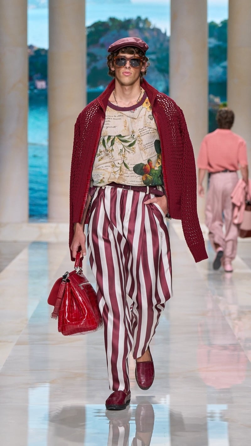 Model in a deep red flat cap and tortoise sunglasses wears a deep red openwork knit cardigan over an oversized Sicilian postcard print tee, paired with wide pleated burgundy and ivory awning-striped silk trousers, deep red loafers, and a red croc-embossed bag