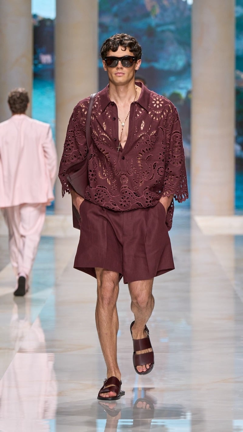 Model in tortoise sunglasses wears an oversized burgundy short-sleeve linen shirt with elaborate floral cutwork embroidery, paired with matching wide pleated burgundy shorts and burgundy slide sandals