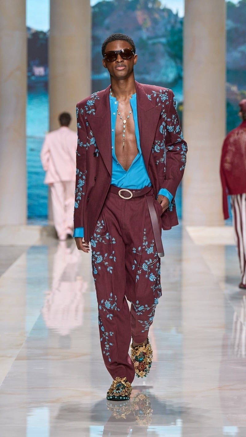 Model in tortoise sunglasses wears an oversized burgundy linen suit densely embroidered with turquoise floral motifs, layered over a turquoise shirt, paired with wide pleated trousers, an ornate buckled belt, and elaborate jeweled brocade slippers