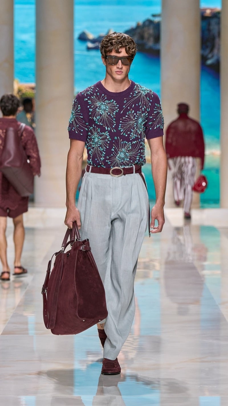 Model in shield sunglasses wears a short-sleeve plum knit crewneck embroidered with turquoise dandelion motifs, paired with wide pleated pale gray linen trousers, a jeweled buckle belt, burgundy loafers, and a burgundy suede travel bag