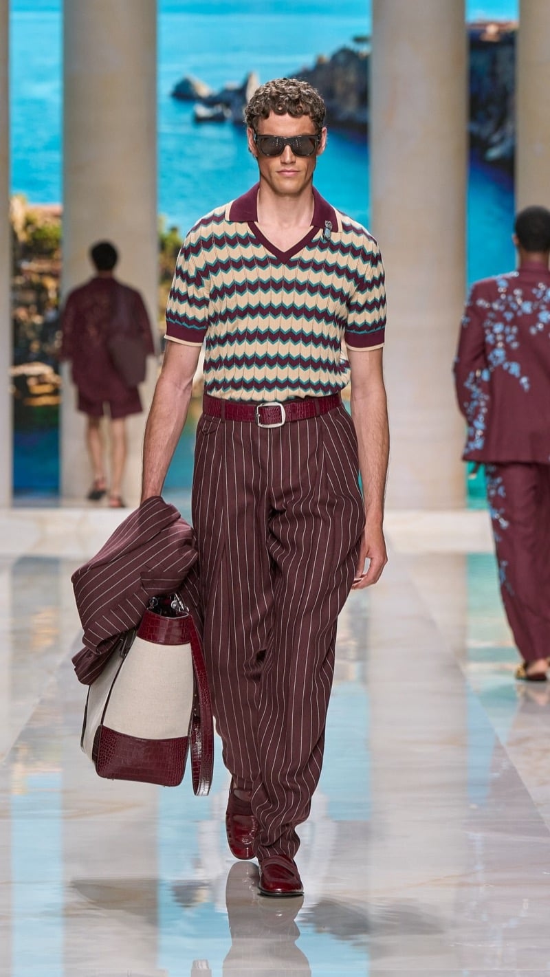 Model in shield sunglasses wears a short-sleeve knit polo with a teal and beige chevron motif, tucked into wide pleated burgundy pinstripe trousers with a red buckled belt, paired with burgundy loafers and a canvas tote with red croc trim