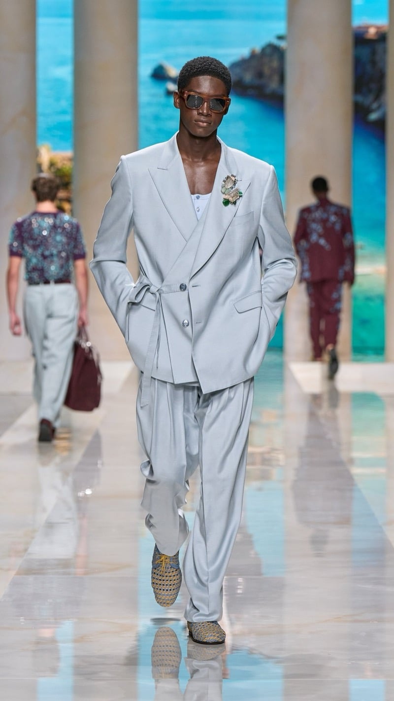 Model in tortoise sunglasses wears an oversized pale gray double-breasted linen suit with peaked lapels and a tied sash, layered over a light tank with a jeweled floral brooch, paired with woven yellow and gray derbies