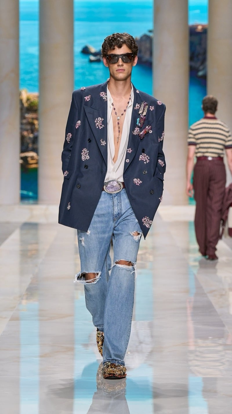 Model in sunglasses wears an oversized deep blue double-breasted linen blazer densely embroidered with pink floral clusters and jeweled brooches, layered over a light shirt, paired with light wash distressed jeans and ornate jeweled brocade slippers
