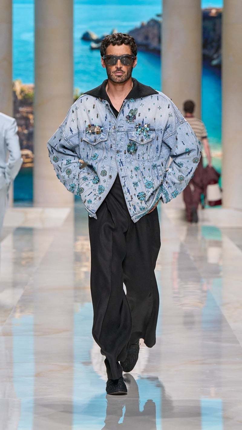 Model in sunglasses wears an oversized light wash denim shirt jacket, densely embroidered with turquoise floral embellishment, paired with wide pleated dark linen trousers and beaded slippers