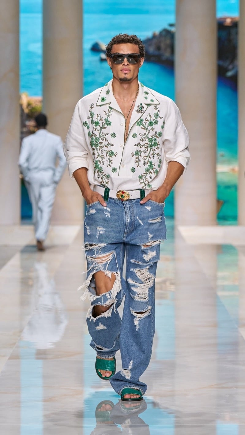 Model in shield sunglasses wears a relaxed light camp-collar shirt embroidered with green floral motifs, paired with heavily distressed light wash jeans, an ornate jeweled belt buckle, and green leather sandals