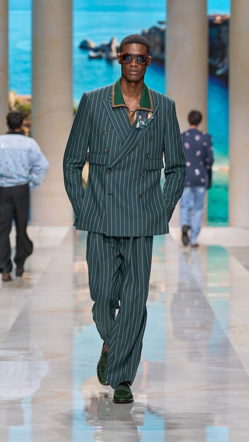 Model in tortoise sunglasses wears an oversized deep green pinstripe linen suit with peaked lapels, layered over a green shirt with contrast trim and a jeweled brooch, paired with wide pleated trousers and green woven derbies