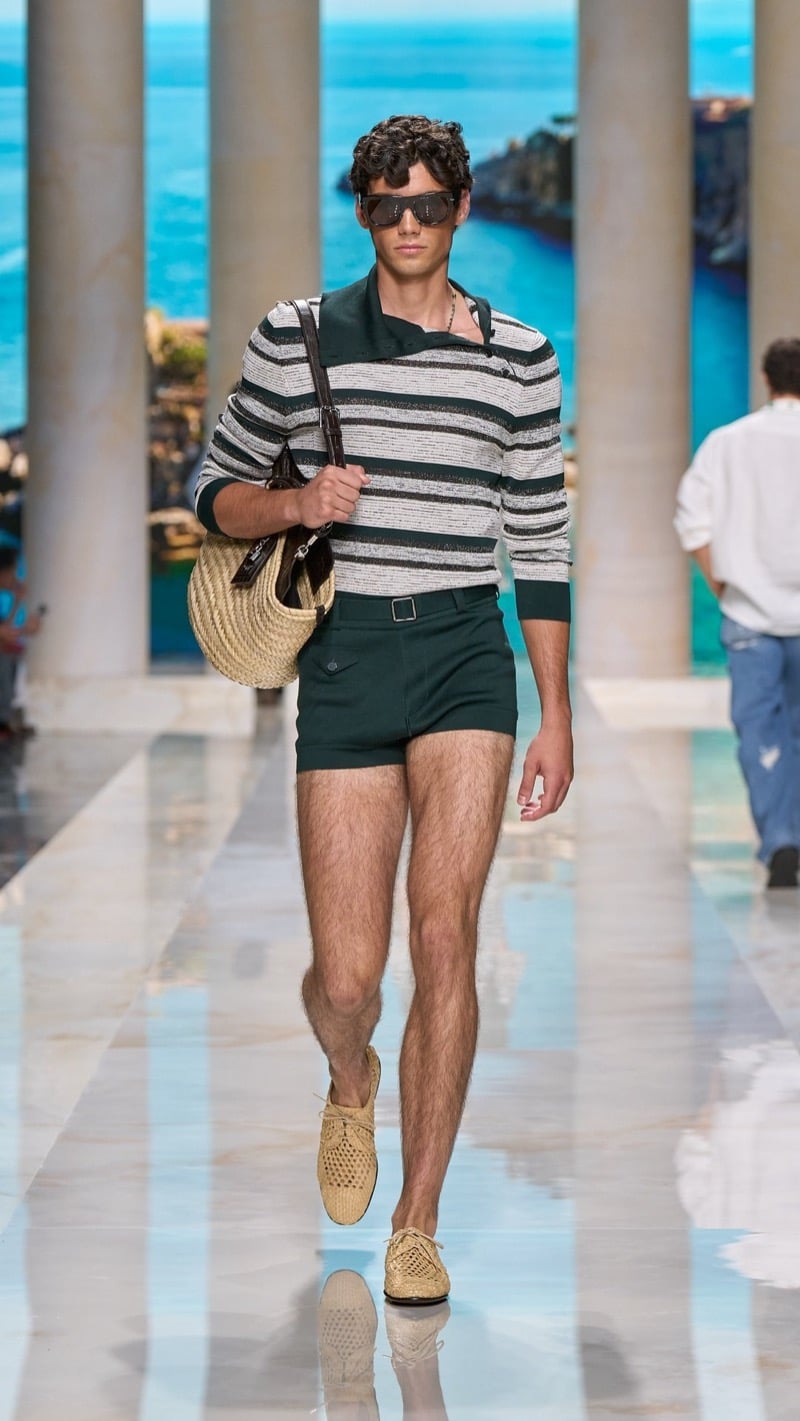 Model in shield sunglasses wears a deep green and ivory striped long-sleeve knit polo tucked into slim belted deep green shorts, paired with crochet raffia derbies and a raffia basket bag with a leather strap