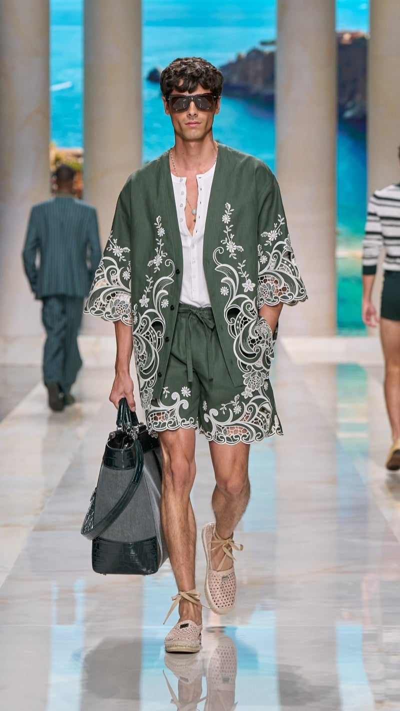 Model in shield sunglasses wears an oversized open deep green linen camp-collar shirt with elaborate ivory floral cutwork embroidery, layered over a light henley, paired with matching deep green drawstring shorts, raffia espadrilles, and a deep green croc-embossed travel bag