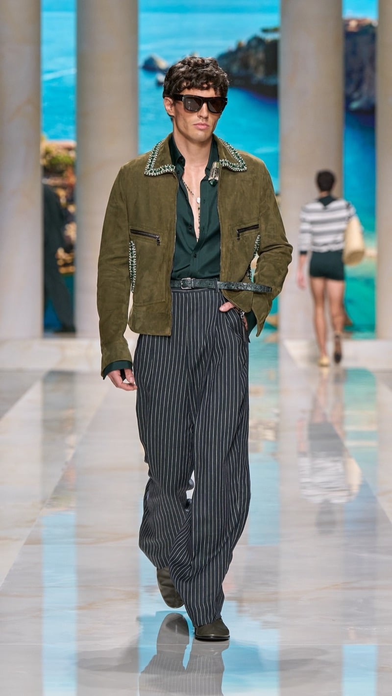 Model in shield sunglasses wears an olive suede jacket with jeweled collar and cuff trim over a deep green shirt, paired with wide pleated dark pinstripe trousers and dark suede shoes