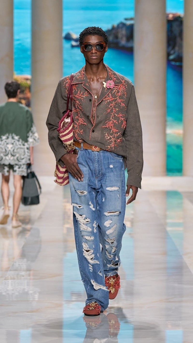 Model in tortoise sunglasses wears a relaxed brown linen camp-collar shirt embroidered with coral motifs, paired with distressed light wash baggy jeans, an ornate buckled belt, and embellished slippers
