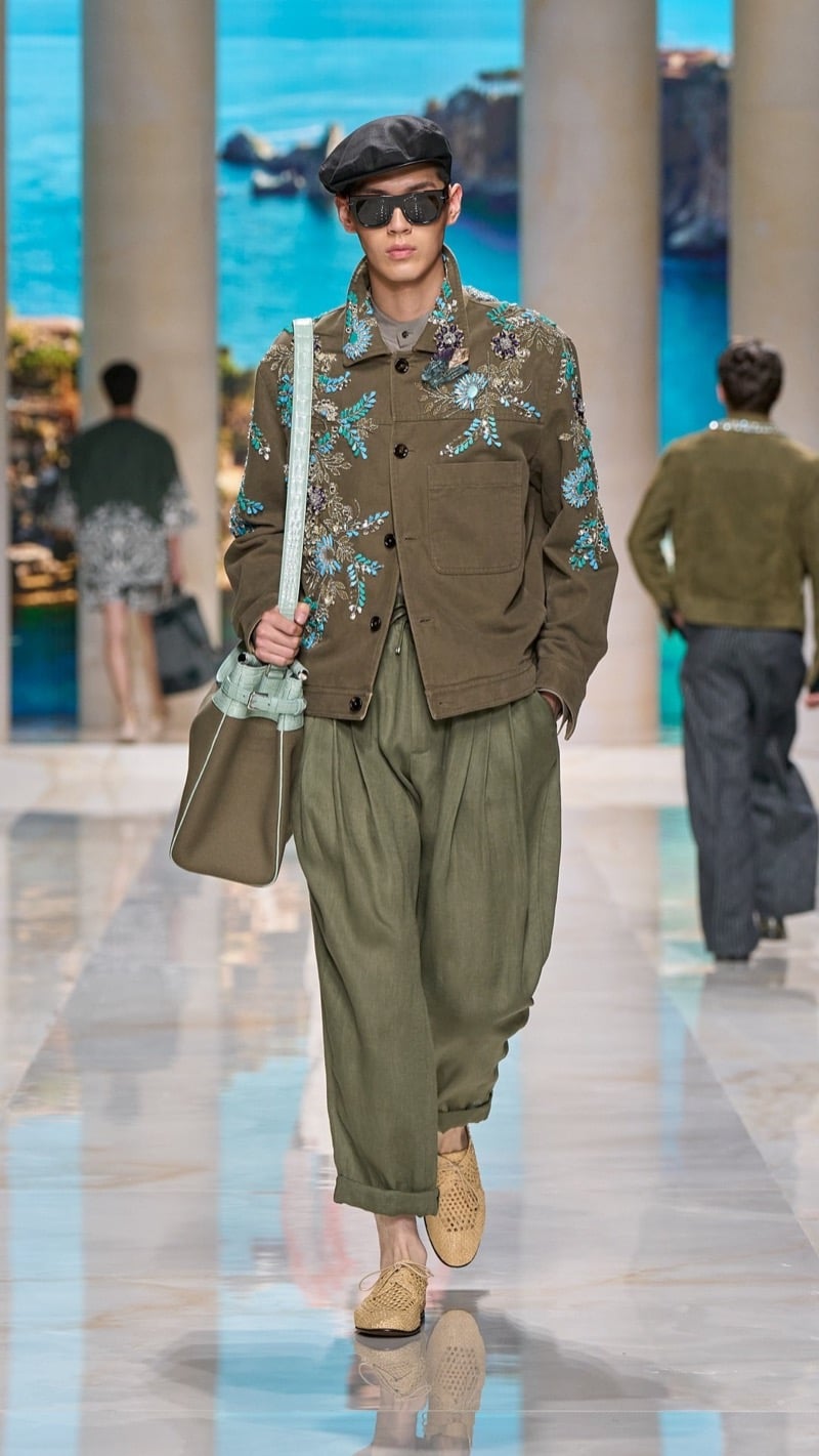 Model in a flat cap and sunglasses wears an olive cotton chore jacket densely embroidered with turquoise floral clusters and jewels, paired with wide pleated cuffed olive linen trousers, raffia derbies, and a sage canvas bag
