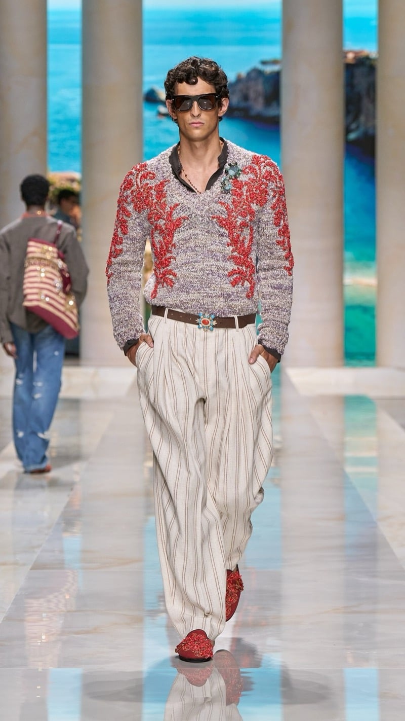 Model in tortoise sunglasses wears a chunky marled V-neck knit sweater intarsia-knit with deep red coral branch motifs, paired with wide pleated ivory pinstripe trousers, a jeweled buckle belt, and embellished red slippers