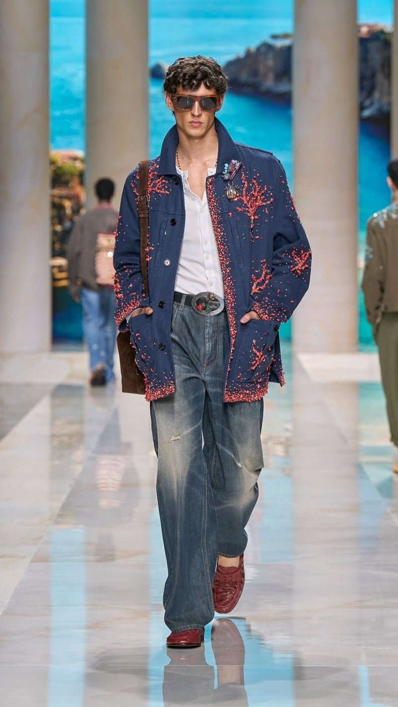 Model in shield sunglasses wears an oversized deep blue cotton chore jacket embroidered with deep red coral motifs over a light shirt, paired with relaxed faded jeans, an ornate buckled belt, and burgundy loafers