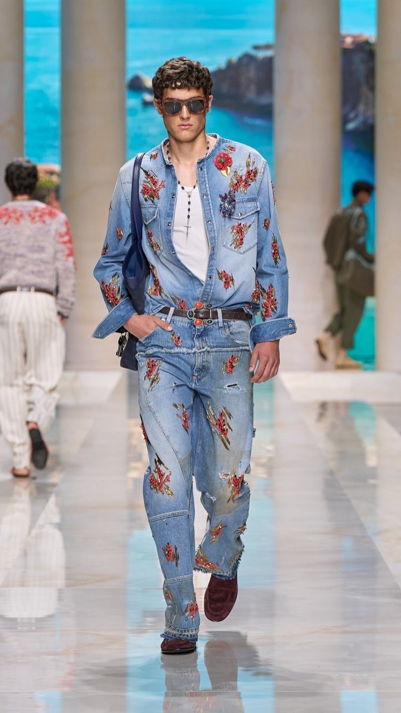 Model in sunglasses wears a light wash denim shirt embroidered with red coral and floral bouquets over a light tank with rosary necklace, paired with matching light wash jeans, a buckled belt, and deep red loafers