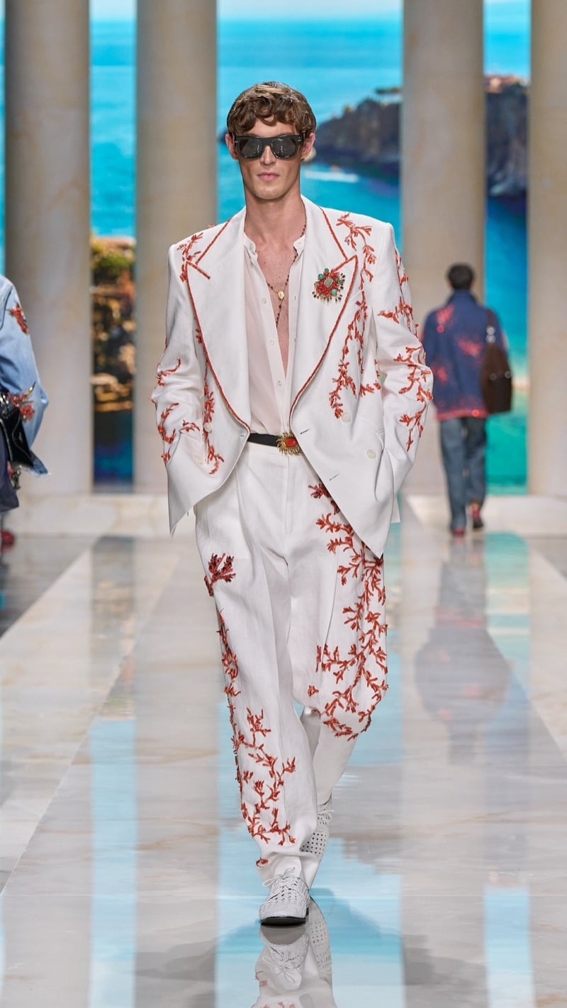 Model in shield sunglasses wears an oversized ivory linen suit densely embroidered with deep red coral branch motifs, layered over a pale silk shirt, paired with wide pleated trousers, a jeweled buckle belt, and woven ivory derbies