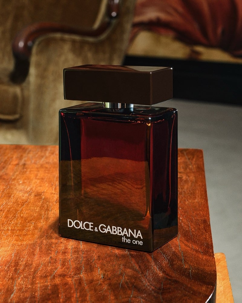 The Dolce & Gabbana The One For Men Parfum bottle in deep red-brown glass with a dark cap sits on a polished wooden side table beside a velvet armchair in a softly lit interior