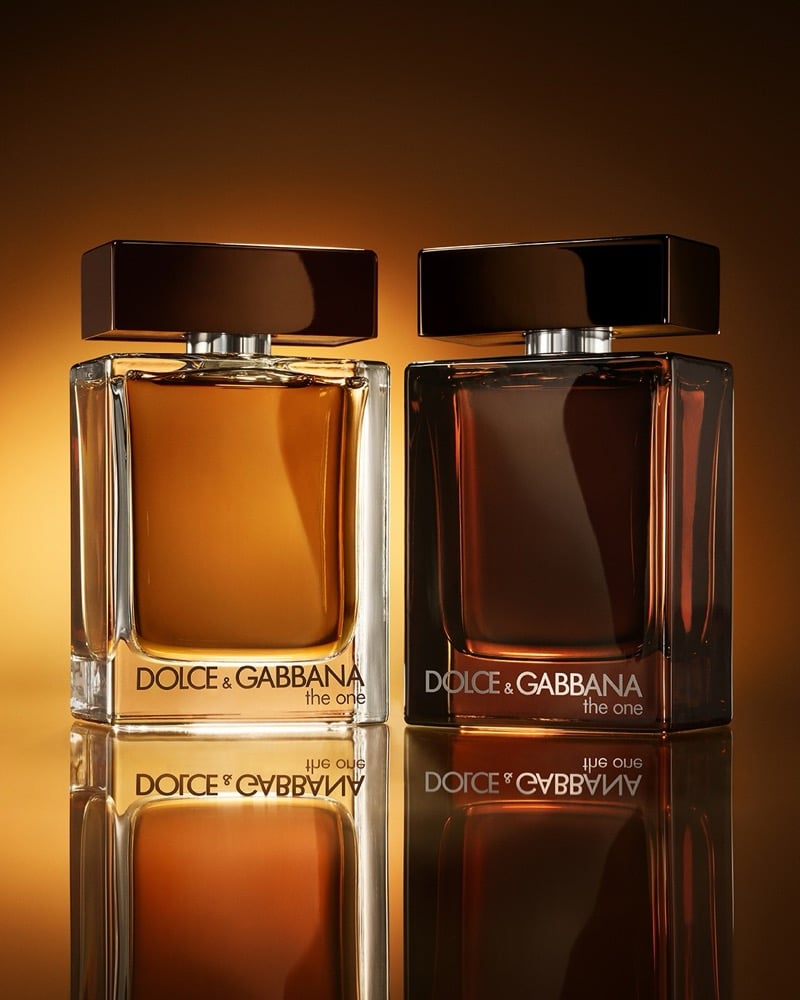 The Dolce & Gabbana The One For Men Eau de Parfum bottle in honey amber sits beside the deeper red-brown The One For Men Parfum, both with dark brown caps, photographed against a warm amber gradient backdrop with mirror reflections