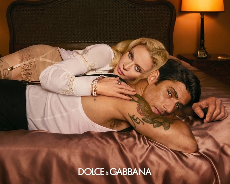 Alberto Guerra in a white ribbed tank top with tattooed arms lies on a satin bedspread beside Madonna