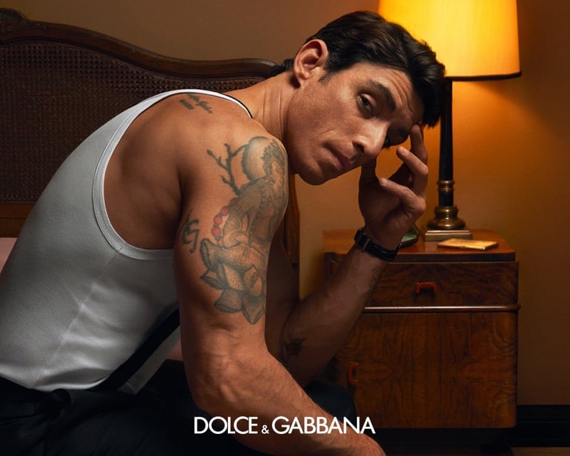 Alberto Guerra sits at the edge of the bed in a white ribbed tank top with black tailored trousers, leaning a tattooed arm against his temple beside a brass table lamp and a wooden nightstand