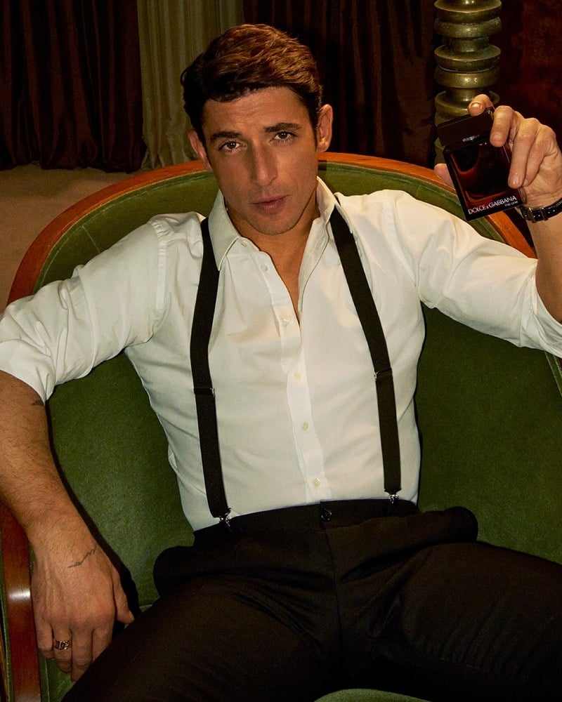Alberto Guerra reclines in a green velvet armchair in a white open-collar shirt with black suspenders and tailored trousers, holding up the dark Dolce & Gabbana The One For Men Parfum bottle beside a brass table lamp