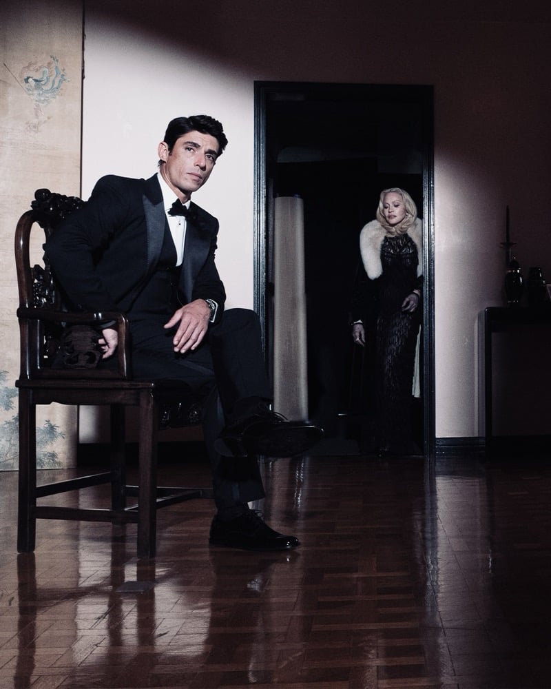 Behind the scenes of the Dolce & Gabbana The One 2026 campaign: Alberto Guerra sits in a carved wooden chair in a black tuxedo and bow tie on a polished parquet floor while Madonna stands in a dark doorway in a black lace gown and pale fur stole