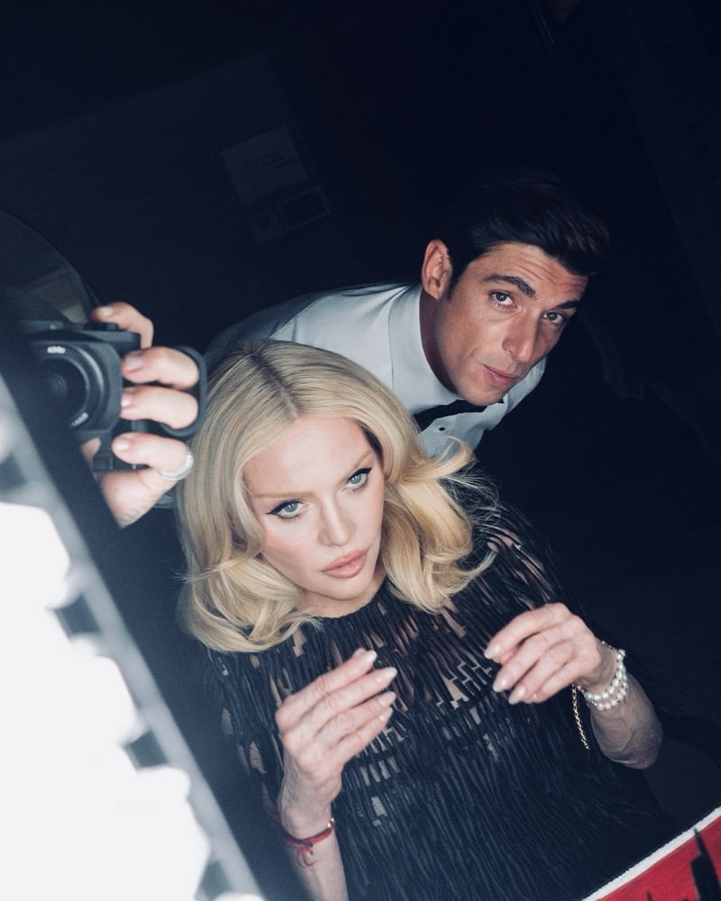 Overhead behind the scenes shot of Alberto Guerra in a white shirt and black bow tie leaning over Madonna's shoulder as she wears a black textured dress, pearl bracelet and a soft platinum blonde wave
