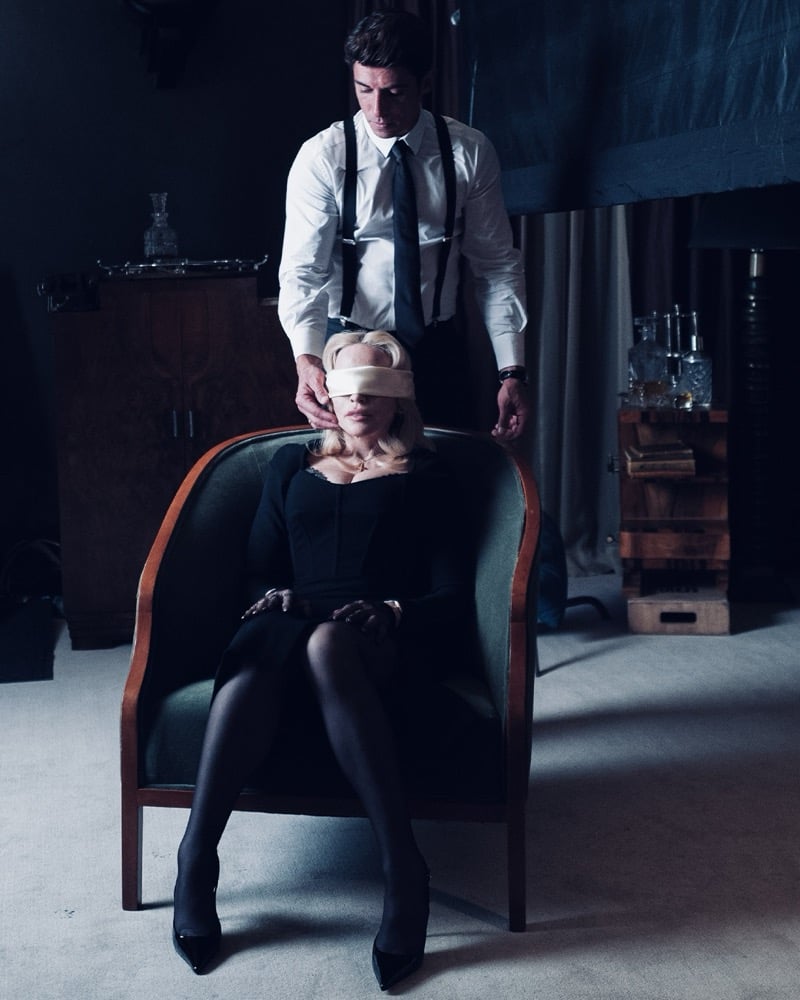 Behind the scenes of the Dolce & Gabbana The One 2026 campaign: Alberto Guerra in a white shirt, black tie and black suspenders ties a cream silk blindfold over Madonna's eyes as she sits in a green armchair in a black dress, black tights and black pointed pumps