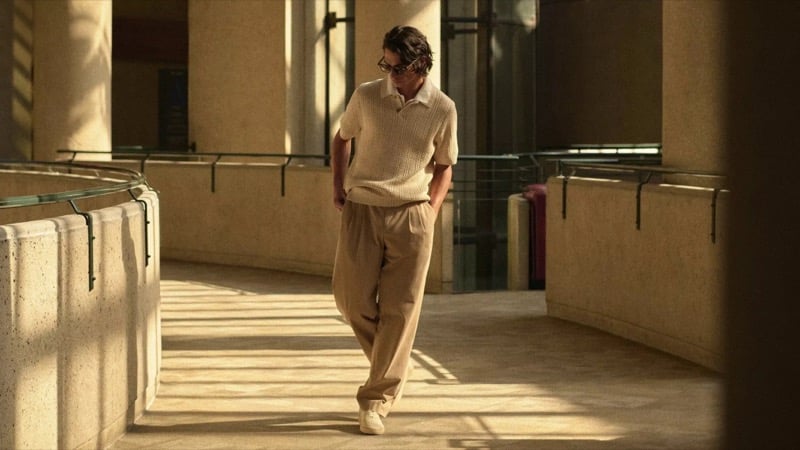 Donovan Wildfong walks along the curved Bonaventure walkway in tortoise glasses, an oatmeal openwork knit polo, and matching pleated trousers with sneakers