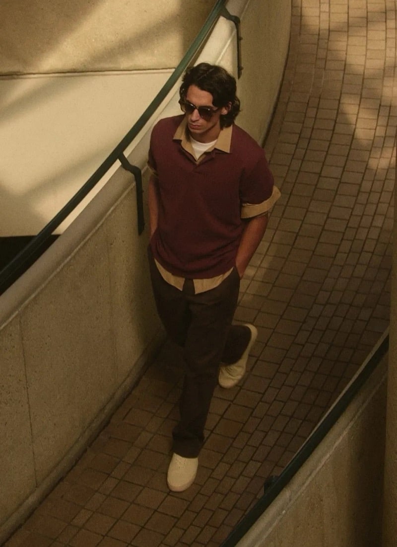 Donovan Wildfong walks a tiled corridor in tortoise sunglasses, a burgundy polo layered over a cream shirt, paired with dark trousers and cream sneakers