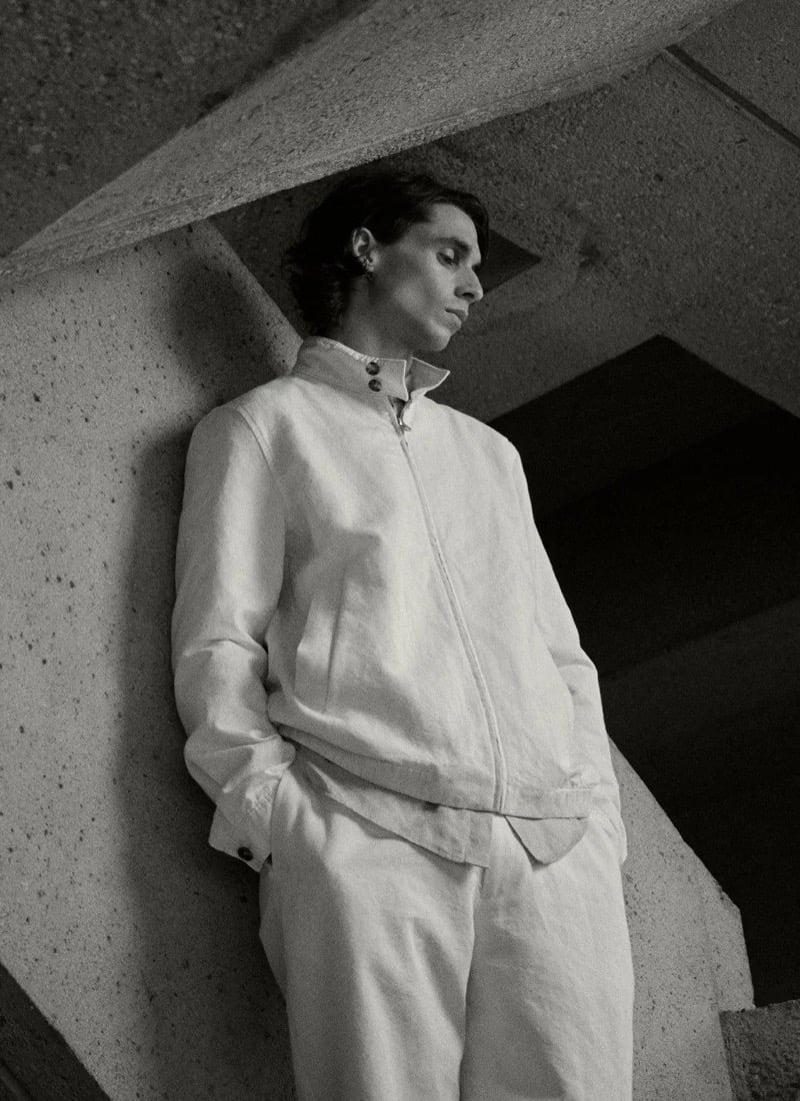 Black-and-white image of Donovan Wildfong leaning against an angular concrete column in a stand-collar zip jacket and matching trousers