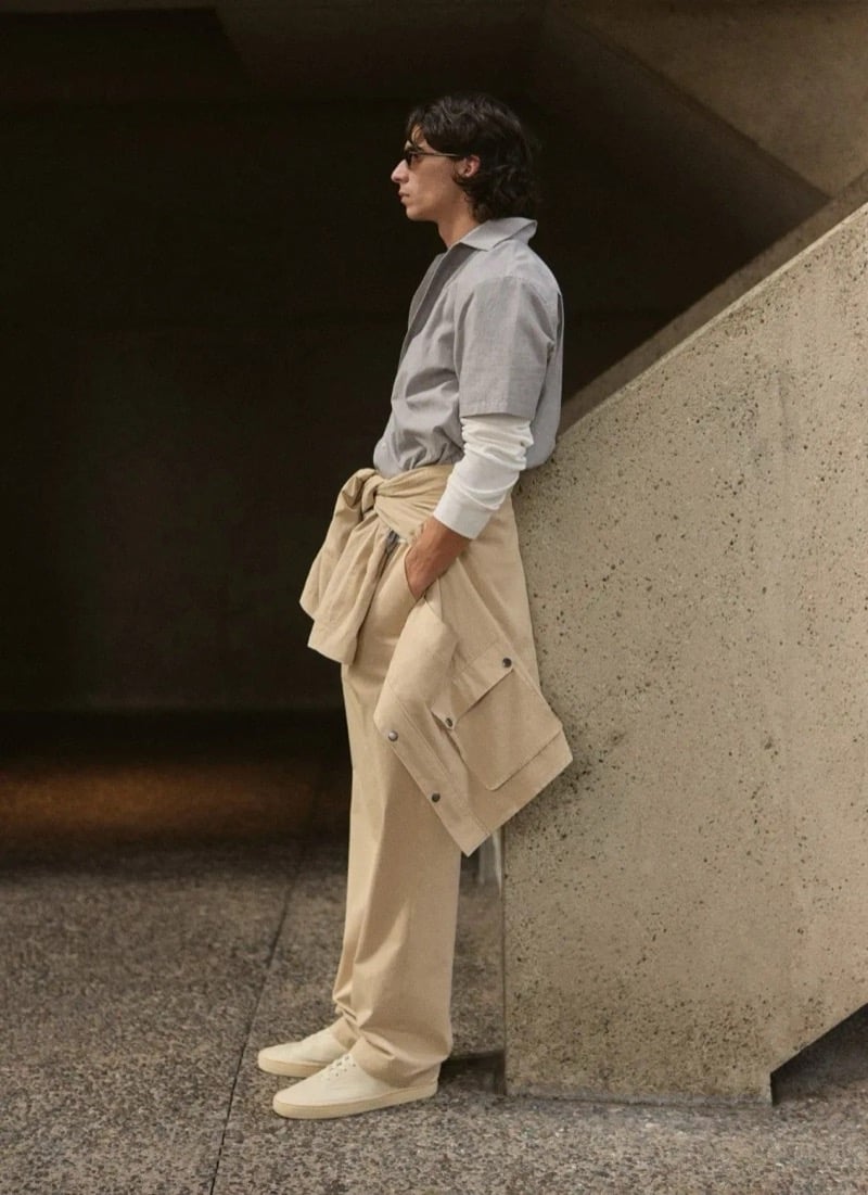 Donovan Wildfong leans against a concrete corner in a slate blue button-down with a tan jacket tied around his waist over wide-leg trousers and cream canvas sneakers