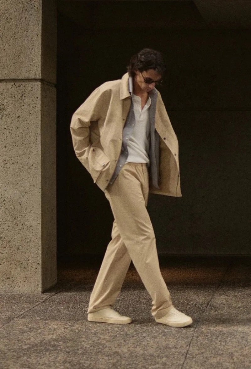 Donovan Wildfong walks beneath a concrete overhang in a tan oversized coach jacket over a layered knit polo and tan trousers with cream sneakers