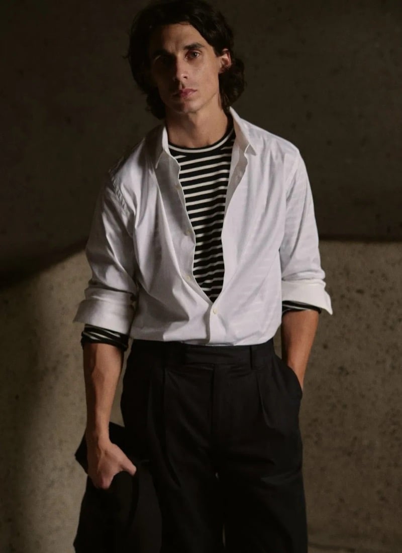 Donovan Wildfong stands against a textured concrete wall in a crisp open button-down layered over a slim striped tee, with dark pleated wide-leg trousers