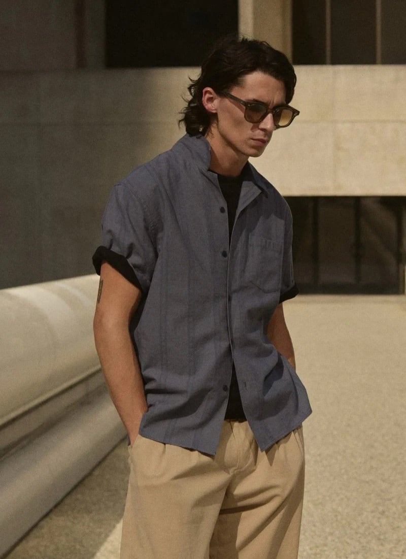 Donovan Wildfong in tortoise sunglasses wears a slate blue short-sleeve camp-collar shirt layered over a dark tee with tan pleated trousers
