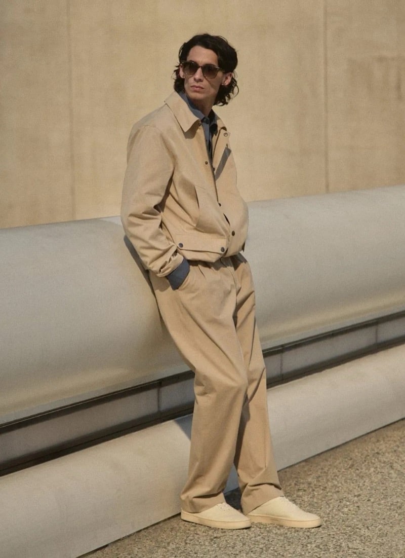 Donovan Wildfong leans against concrete in tortoise sunglasses, a tan cotton harrington jacket over a shirt and tonal pleated trousers, finished with cream canvas sneakers