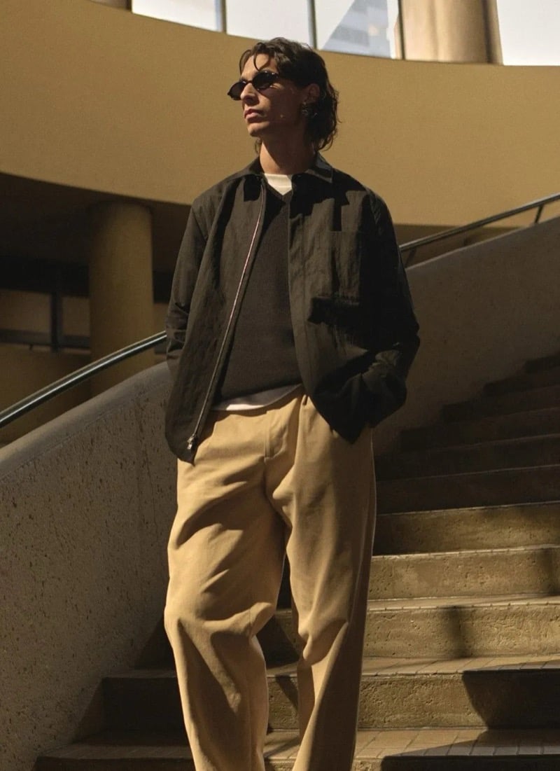 Donovan Wildfong in slim sunglasses wears a dark abstract-print zip jacket over a brown crewneck sweater and tan pleated trousers on the Bonaventure's curved staircase