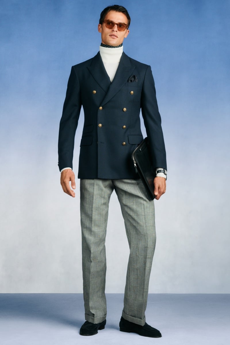 Model Parker Van Noord in tortoise sunglasses wears a navy double-breasted blazer with gold buttons over a cream turtleneck and printed neckerchief, paired with gray Prince of Wales check trousers, black suede loafers, and a black leather portfolio